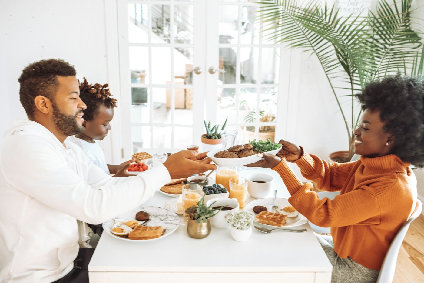 A mother, father and son share a meal at a sunny dining table, passing a plate of food
