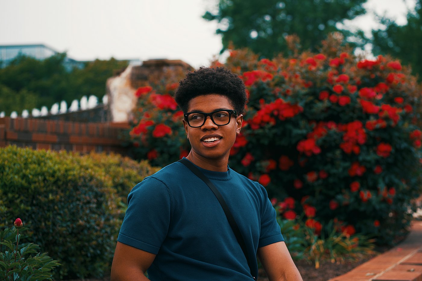 A smiling teen with glasses and a shoulder bag looks off to the side in front of red flowering bushes