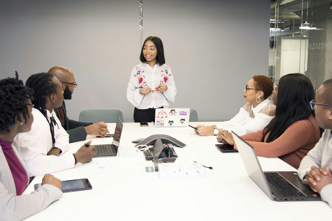 A young woman stands at the head of a conference table leading a meeting while colleagues listen