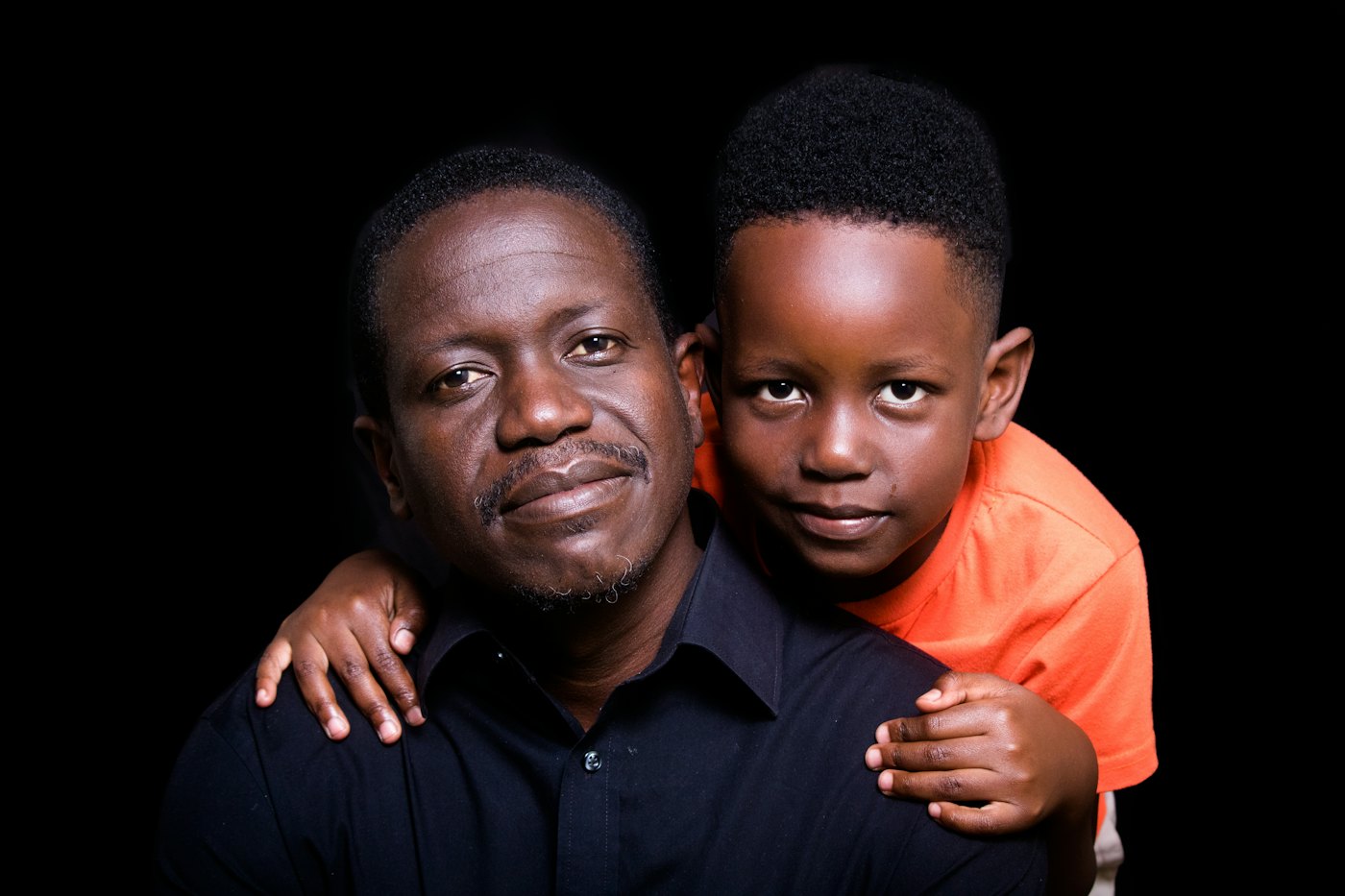 A boy hugs a smiling man from behind in a warm portrait