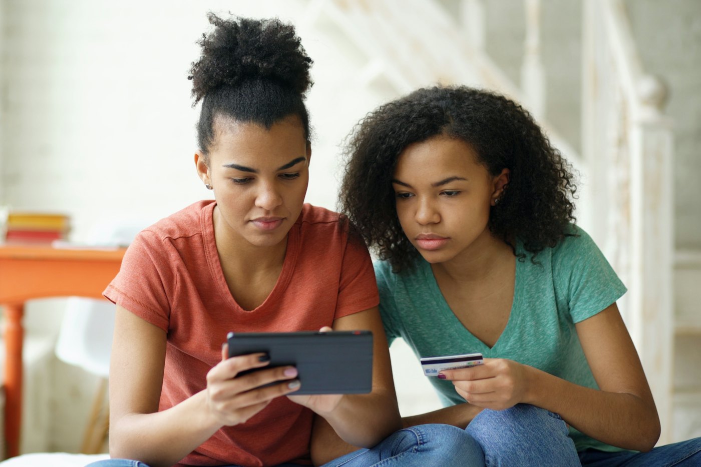 Two teen girls look at a tablet together, one holding a debit card