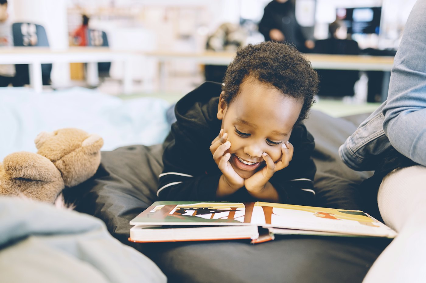 A young boy grins while reading a picture book, chin in his hands, on a beanbag